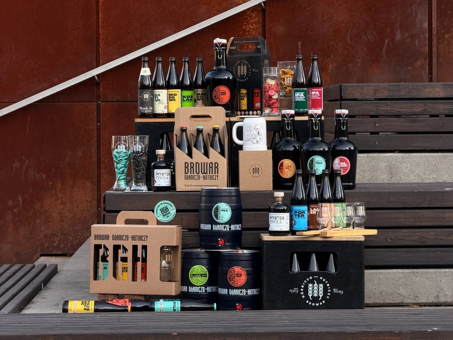 BGH beers, three-packs, kegs and glassware arranged on wooden steps by a rusted steel wall